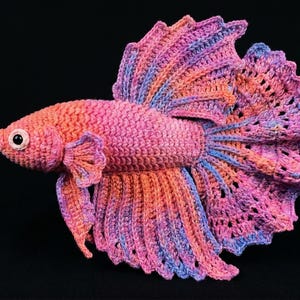 May include: A handmade crochet Betta fish in shades of pink, orange, and blue. The fish has detailed fins and a textured body, with a black background. The fish is a decorative item.