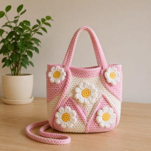 May include: A handmade crochet tote bag in pink and cream, adorned with white and yellow daisy appliqués. The bag features a shoulder strap and two handles. The bag is sitting on a wooden surface.