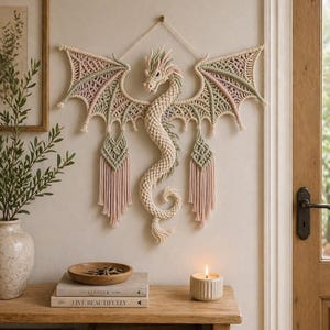 Könnte beinhalten: Ein Wandbehang aus Makramee, der einen Drachen mit rosa und grünen Flügeln darstellt. Der Drachenkörper ist cremefarben, mit einem langen, gewundenen Schwanz. Die Flügel sind mit komplizierten Knoten versehen.