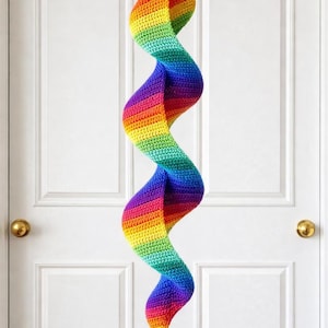 May include: A handmade, rainbow-colored crochet spiral hanging decoration. The spiral is suspended from a teal string and features a gradient of colors, including red, orange, yellow, green, blue, and purple. A small, white ball hangs at the bottom.