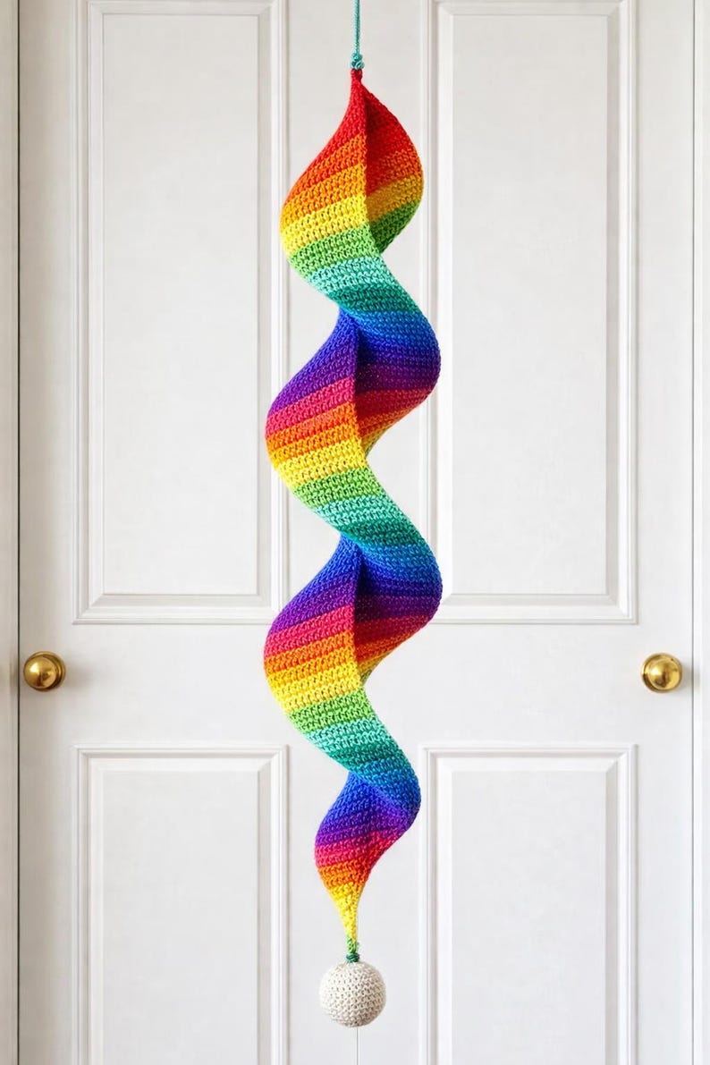 May include: A handmade, rainbow-colored crochet spiral hanging decoration. The spiral is suspended from a teal string and features a gradient of colors, including red, orange, yellow, green, blue, and purple. A small, white ball hangs at the bottom.