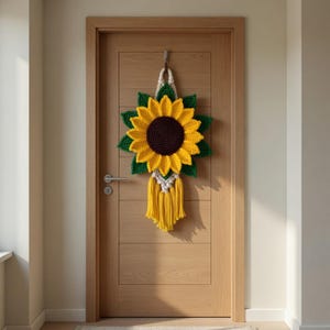 May include: A handmade crochet sunflower door hanging. The sunflower has yellow petals, a brown center, and green leaves. A white macrame cord and yellow fringe hang below the flower. The door is made of wood.