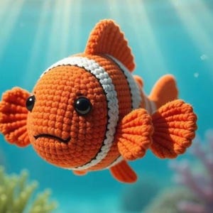 May include: A crocheted orange and white clownfish with black eyes and a slightly downturned mouth. The fish has orange fins and is set against a blurred blue background, evoking an underwater scene. The fish is a handmade item.