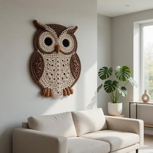 Op de afbeelding: Een grote, handgemaakte gehaakte uil voor aan de muur. De uil heeft een bruin en crèmekleurig kleurenschema met zwarte ogen en een gedetailleerd, gestructureerd ontwerp. De uil is aan een lichte muur bevestigd en voegt een vleugje rustieke charme toe.