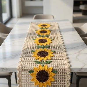 Puede incluir: Camino de mesa de ganchillo color crema con un patrón repetido de girasoles. Cada girasol tiene pétalos amarillos, un centro marrón y hojas verdes. El camino de mesa está colocado sobre una mesa de mármol blanco.