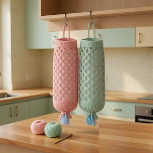 Può includere: Due sacchetti per prodotti all'uncinetto, uno rosa e uno verde, sono appesi a dei ganci. Le borse hanno un motivo a rombi e un manico. Due gomitoli di lana, rosa e verde, e un uncinetto sono sul bancone. Le borse sono lunghe circa 30 cm.