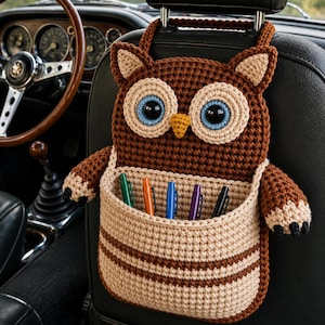 Può includere: Un organizer a forma di gufo realizzato all'uncinetto nei colori marrone e beige, appeso al poggiatesta di un'auto. Il gufo presenta grandi occhi blu e bianchi, un becco giallo e due braccia laterali. Una tasca beige frontale contiene cinque penne.