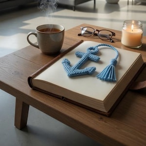 May include: A blue knitted anchor bookmark with a tassel rests on an open book with a brown leather cover. A cup of tea, glasses, and a lit candle sit on a wooden table, creating a cozy reading scene.