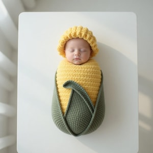 Puede incluir: Un bebé recién nacido con un gorro amarillo de ganchillo y envuelto en un disfraz de maíz de ganchillo amarillo y verde. El bebé está durmiendo plácidamente sobre una superficie blanca. El disfraz se asemeja a una mazorca de maíz.