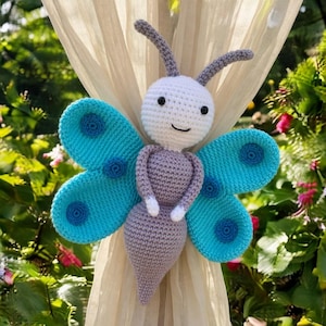 May include: A handmade crochet butterfly in shades of white, teal, and gray. The butterfly has large teal wings with darker blue circles, a white head, and a gray body. The butterfly is designed to be a curtain tieback.