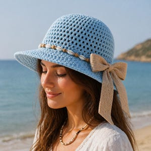 Puede incluir: Un sombrero de ganchillo azul claro con un ala pequeña, adornado con conchas marinas y un lazo beige. El sombrero tiene una banda decorativa de pequeñas conchas y cuentas de madera. Un accesorio elegante para los días soleados.
