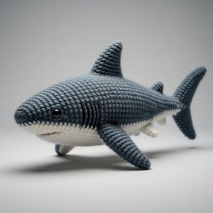 Puede incluir: Un juguete de tiburón tejido a ganchillo en azul y blanco. El tiburón tiene un cuerpo texturizado, una aleta dorsal y una aleta caudal. La parte inferior y la boca son blancas, con ojos negros. Un juguete suave.