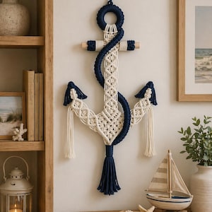 Puede incluir: Un colgante de pared de ancla de macramé de temática náutica. El ancla presenta un cuerpo de color crema con detalles y borlas azul marino. La decoración de la pared se exhibe en una pared neutra, con otros elementos de decoración náutica en el fondo.