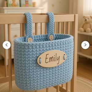 May include: A light blue, handmade crochet basket with wooden button accents and a wooden name tag that reads "Emily." The basket hangs from a wooden crib. The nursery decor includes books and a teddy bear.