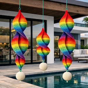 May include: Three rainbow-coloured crochet wind chimes hanging outdoors. Each chime has a spiral design with red, orange, yellow, green, blue, and purple hues. A white crochet ball hangs at the bottom of each chime. The chimes are suspended by teal-coloured cords.