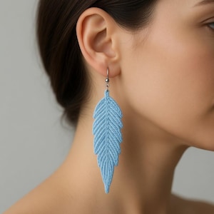 May include: A single, light blue feather earring with a silver hook. The earring is made of a woven material, with detailed lines creating the feather's shape. The earring is hanging from the ear of a person.