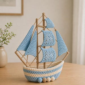 Könnte beinhalten: Ein handgefertigtes Häkelschiff in Hellblau, Beige und Braun. Das Boot hat detaillierte Segel und einen gestreiften Rumpf mit dekorativen Kugeln am Boden. Ein charmantes Deko-Objekt zum Thema Seefahrt.