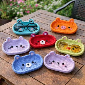 May include: A collection of handmade crochet eyeglass holders in various colors, including blue, orange, red, and green. Each holder is shaped like an animal face, such as a frog, pig, or bear, and features embroidered details.