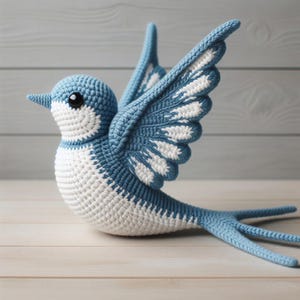 Pode incluir: Pássaro de crochê azul e branco com asas abertas. O pássaro feito à mão apresenta um design detalhado com um corpo azul, detalhes brancos e uma cauda longa e pontiaguda. O pássaro é exibido em uma superfície de madeira clara.