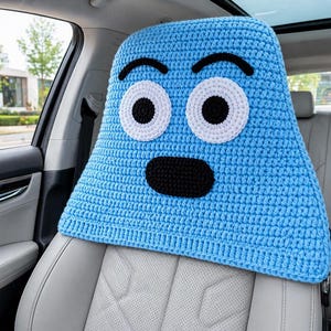 May include: A blue crocheted car seat headrest cover, designed as a cartoon face. It has large white eyes with black pupils, a black mouth, and black eyebrows. The cover is placed on a light grey car seat inside a car.