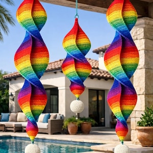 May include: Three rainbow-colored, spiral-shaped crochet ornaments hanging outdoors. Each ornament features a vibrant spectrum of colors and is suspended by a teal string. A white, crocheted ball hangs at the bottom of each spiral.