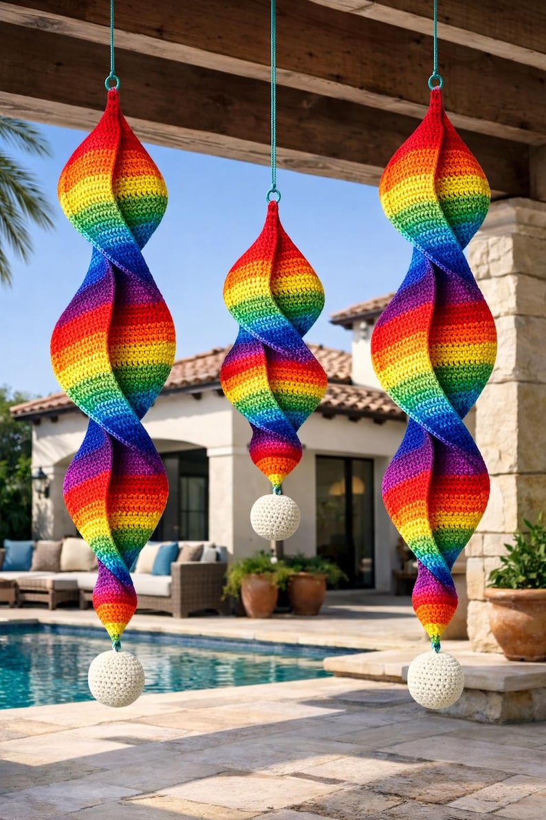 May include: Three rainbow-colored, spiral-shaped crochet ornaments hanging outdoors. Each ornament features a vibrant spectrum of colors and is suspended by a teal string. A white, crocheted ball hangs at the bottom of each spiral.