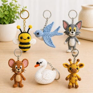 May include: A collection of handmade crochet keychains featuring various animal designs. Includes a yellow and black bee, a blue bird, a gray cat, a brown mouse, a white swan, and a yellow giraffe. Each keychain has a metal ring for attaching to keys or bags.
