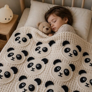 May include: A cream-colored, crocheted blanket featuring multiple panda faces with black and white details. A child is sleeping under the blanket with a teddy bear. The blanket is on a bed with a wooden frame.