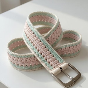 May include: A crocheted belt in pastel colors. The belt features a repeating pattern of pink and light green sections, bordered by a cream-colored yarn. A silver-colored metal buckle secures the belt. The belt is displayed on a white surface.