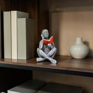 May include: A decorative figurine of a person reading a book, with a black and white pattern. The book is open and coloured orange. The figurine is sitting cross-legged. A white vase is to the right.
