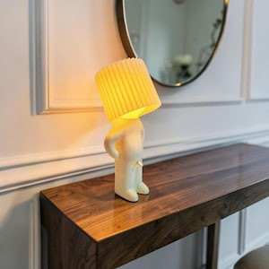 May include: A table lamp featuring a cream-colored, human-shaped base with a standing posture. The lamp head is a cylindrical, pleated, off-white lampshade that emits a warm, soft yellow glow, illuminating the figure from above.