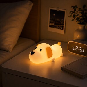 May include: A soft, glowing silicone night light shaped like a puppy. The lamp features a smooth, rounded body with a warm yellow light, dark brown ears, a black nose, and black eyes. It is positioned horizontally, emitting a gentle illumination.