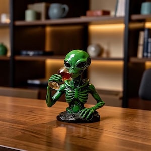 May include: A green alien bust figurine with a skeletal chest structure, large black eyes, and an elongated head. The alien is holding a clear glass filled with a dark amber liquid up to its mouth, featuring a detailed textured base and glossy finish.
