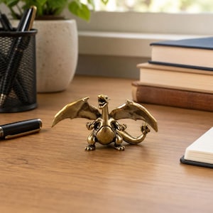May include: A small, metallic, brass-coloured dragon figurine with outstretched wings, a wide-open mouth, and a curled tail. The creature stands in a bipedal pose with its arms raised, featuring a smooth, reflective surface and detailed sculpted features.