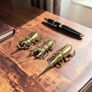 May include: Three brass beetle figurines with detailed exoskeletons and legs are arranged on a wooden surface. A black pen with silver accents rests nearby. The beetles vary in design, showcasing different types of insect anatomy.