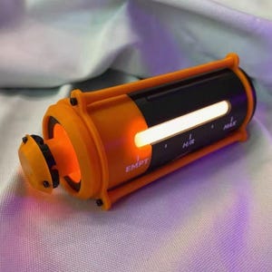 Puede incluir: Un dispositivo cilíndrico de color naranja y negro con una barra de luz horizontal blanca brillante. La carcasa naranja presenta el texto impreso EMPT, H/R y MAX en el lateral. Una pequeña tapa naranja está unida a un extremo del cuerpo principal.