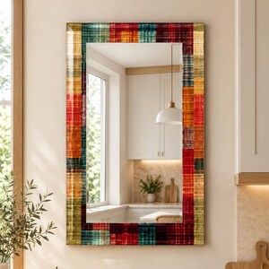 May include: A rectangular wall mirror featuring a frame with a multi-colored plaid pattern in shades of red, orange, yellow, and teal. The glass surface is clear and reflective, showing a kitchen interior with white cabinets and a window.