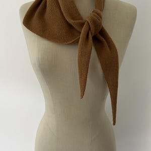 May include: A solid brown knitted scarf tied in a simple knot around the neck of a mannequin. The scarf features a fine, uniform knit texture and pointed ends, draping symmetrically across the chest area with a consistent warm brown color throughout.