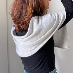 May include: A light gray hooded shawl. The shawl is draped over a black top and has a soft, textured appearance. The hood is folded over the shoulders, creating a layered effect. The sleeves are wide and flowing.