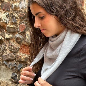 May include: A light grey triangular knit scarf worn around the neck, featuring a soft, fine-gauge texture. The scarf is draped in a simple fold, contrasting against a plain black long-sleeved top, showcasing a clean and neutral aesthetic.