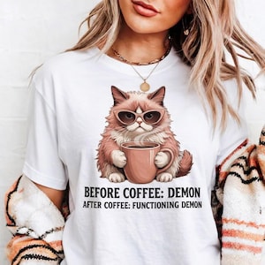 May include: White t-shirt featuring a cartoon cat wearing sunglasses and holding a coffee mug. The cat is pink and brown with a grumpy expression. Text on the shirt reads "BEFORE COFFEE: DEMON AFTER COFFEE: FUNCTIONING DEMON."