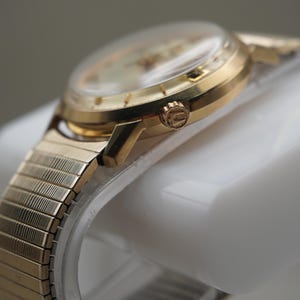 Op de afbeelding: Een goudkleurig polshorloge met een ronde kast en een flexibele, rekbare metalen schakelband. De zijkant van de horlogekast toont een kleine, getextureerde opwindkroon met een zichtbaar logo, terwijl de wijzerplaat is bedekt door een helder kristal.