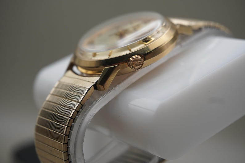 Op de afbeelding: Een goudkleurig polshorloge met een ronde kast en een flexibele, rekbare metalen schakelband. De zijkant van de horlogekast toont een kleine, getextureerde opwindkroon met een zichtbaar logo, terwijl de wijzerplaat is bedekt door een helder kristal.