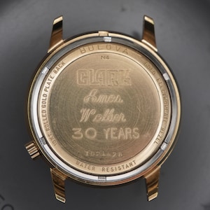 Op de afbeelding: Een goudkleurige Bulova horlogekastbodem met een gravure die CLARK, Ames, Walker, 30 YEARS leest. De metalen behuizing bevat tekst met 10K ROLLED GOLD PLATE BACK, GOLD ELECTROPLATE BEZEL, N4 en WATER RESISTANT, met een kleine kroon aan de zijkant.