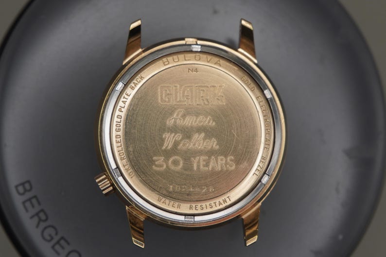 Op de afbeelding: Een goudkleurige Bulova horlogekastbodem met een gravure die CLARK, Ames, Walker, 30 YEARS leest. De metalen behuizing bevat tekst met 10K ROLLED GOLD PLATE BACK, GOLD ELECTROPLATE BEZEL, N4 en WATER RESISTANT, met een kleine kroon aan de zijkant.