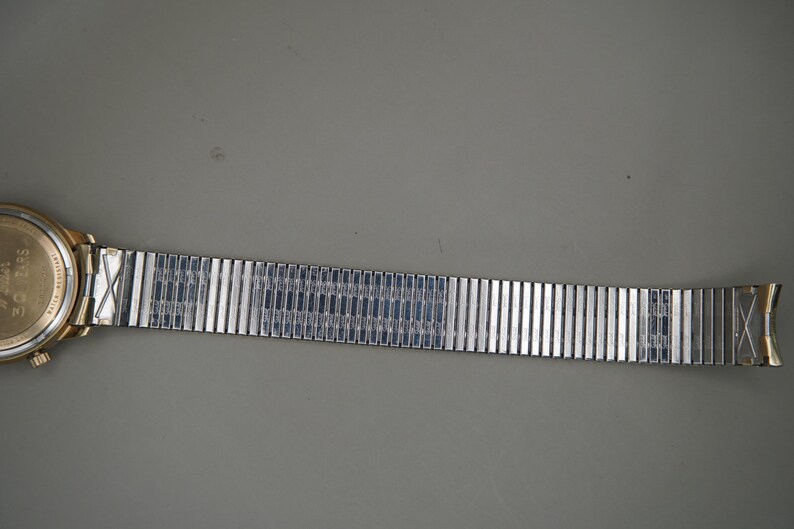 Op de afbeelding: Een goudkleurig polshorloge met een ronde kast, een metalen achterkant en een flexibele, rekbare zilverkleurige metalen schakelband. De horlogeband bestaat uit talloze rechthoekige segmenten die met interne veren met elkaar verbonden zijn.