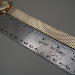 Op de afbeelding: Een goudkleurig polshorloge met een flexibele metalen expansieband naast een metalen liniaal. De wijzerplaat is rond met een goudkleurige afwerking. De liniaal toont de lengte van de horlogeband die zich uitstrekt van nul tot ongeveer 18 cm.
