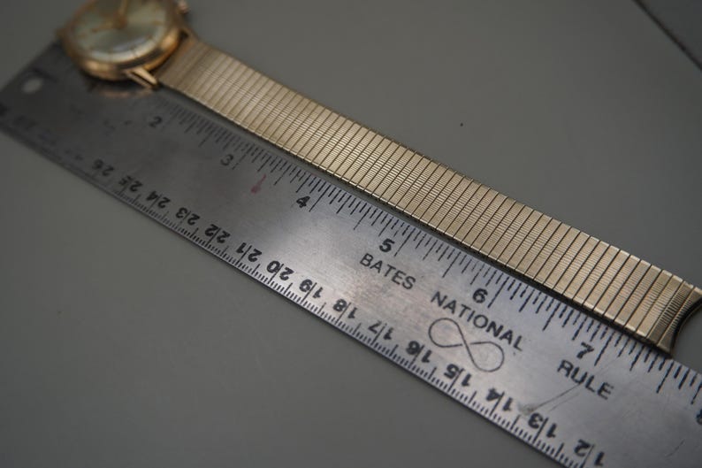 Op de afbeelding: Een goudkleurig polshorloge met een flexibele metalen expansieband naast een metalen liniaal. De wijzerplaat is rond met een goudkleurige afwerking. De liniaal toont de lengte van de horlogeband die zich uitstrekt van nul tot ongeveer 18 cm.