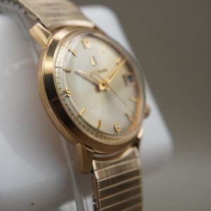 Op de afbeelding: Een goudkleurig polshorloge met een ronde wijzerplaat, een lichtgekleurde wijzerplaat en goudkleurige uurmarkeringen. Het horloge heeft een metalen rekbare band met een textuur en een klein datumvenster op de drie-uur-positie.