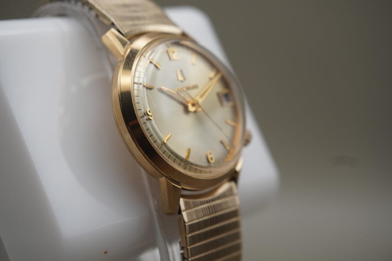 Op de afbeelding: Een goudkleurig polshorloge met een ronde wijzerplaat, een lichtgekleurde wijzerplaat en goudkleurige uurmarkeringen. Het horloge heeft een metalen rekbare band met een textuur en een klein datumvenster op de drie-uur-positie.
